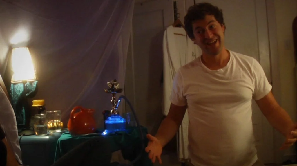 Humpday (2009) movie still: medium — A dimly lit room contains a glowing blue hookah device on a table. A man stands in front of the camera with his…
