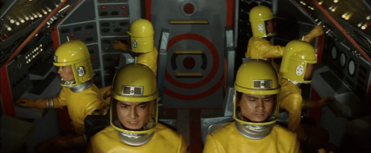 Destroy All Monsters (1968) movie still: medium — Five crew members in bright yellow uniforms sit in a cramped, multi-station spaceship control…