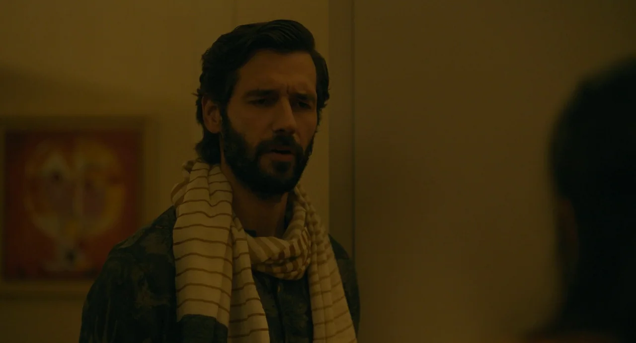 Confess, Fletch (2022) movie still: medium — A dimly lit room features a man standing against a wall. The man wears a patterned shirt and scarf…