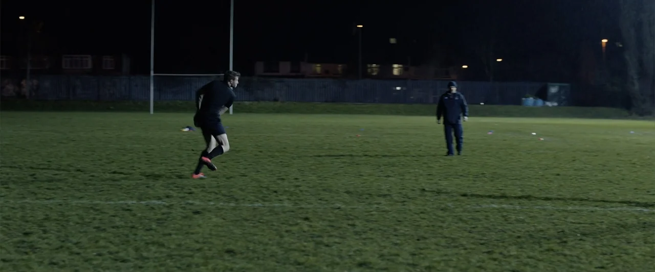 The Thorn On His Side (2014) movie still: wide — An empty nighttime rugby practice field is illuminated by overhead perimeter stadium lights. An…