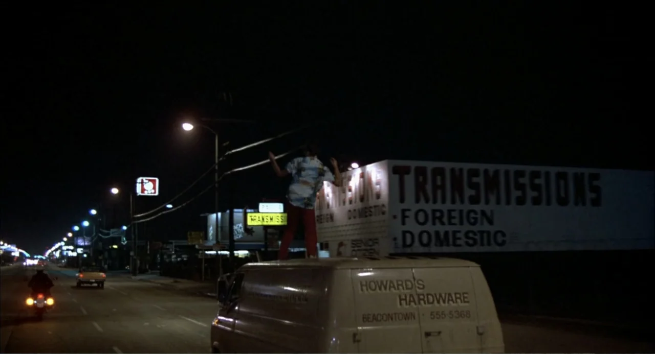 Teen Wolf (1985) movie still: wide — A nighttime view of a commercial street with a large transmission repair building exterior. A man in a…