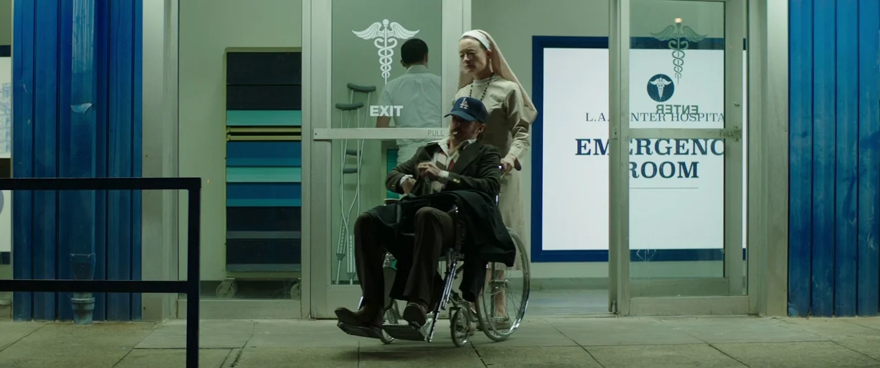 The Nice Guys (2016) movie still: fullbody — The entrance to an emergency room is framed by bright blue corrugated metal panels. A woman dressed as…
