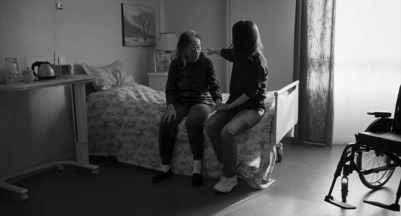 Paris, 13th District (2021) movie still: fullbody — A clinical patient room features a single bed near a large window. Two women sit together on the…