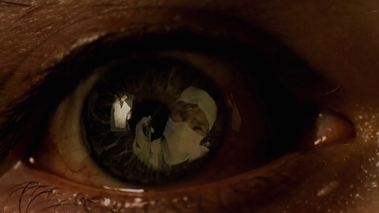Jason X (2001) movie still: closeup — A close-up view shows the iris and pupil of a human eye. A medical professional wearing a surgical cap and…
