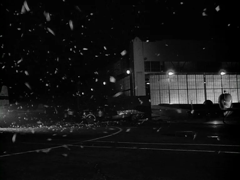 The Killing (1956) movie still: wide — A dark airfield at night features a large hangar with illuminated windows. Confetti or paper drifts through…
