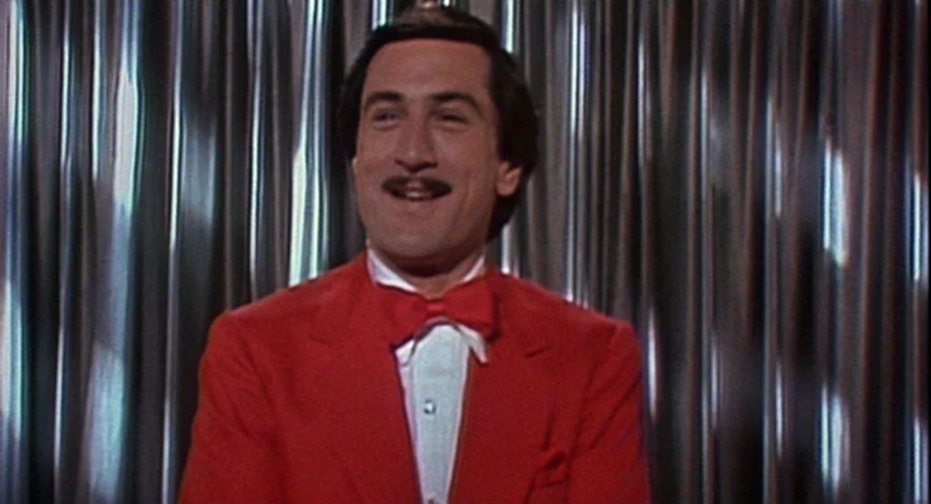 The King Of Comedy (1982) movie still: medium — A man in a red formal suit stands before a reflective metallic curtain. He smiles directly at the…