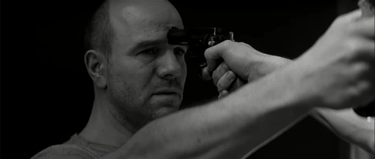 13 Tzameti (2006) movie still: closeup — A man stands in a dark environment with a black background. A handgun is held against the man's forehead by…
