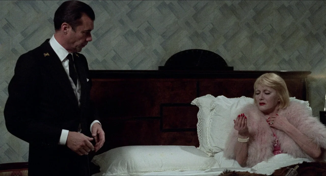 The Night Porter (1974) movie still: medium — A hotel room features a dark wood headboard and textured wallpaper. A man in a suit looks toward a…