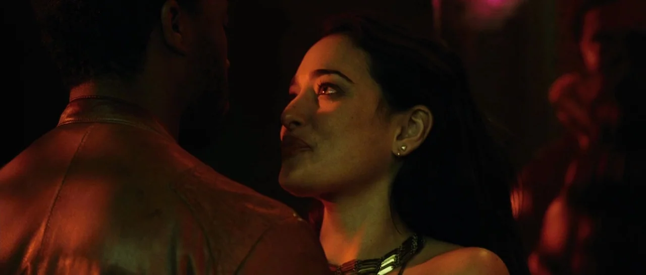 Message From The King (2016) movie still: medium — A dimly lit club interior with red neon lighting illuminating the two subjects. A woman looks up…