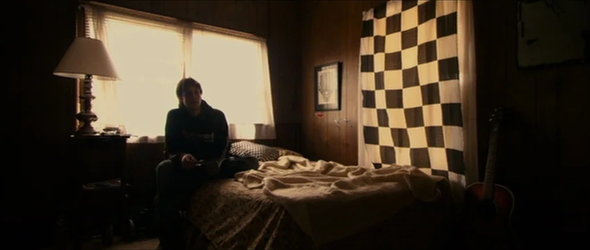 Bubble (2005) movie still: medium — A dark bedroom features wood-paneled walls and a window with bright daylight streaming through. A young man sits…