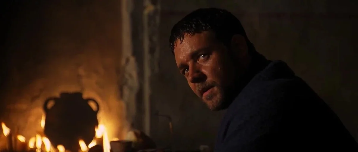 Gladiator (2000) movie still: closeup — A dark stone room is lit primarily by an open hearth fire. A man with short dark hair looks toward the…