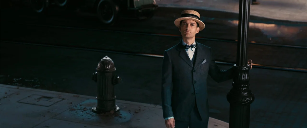 The Great Gatsby (2013) movie still: medium — A nighttime sidewalk at an intersection features a metal lamppost and a fire hydrant. A man in a 1920s…