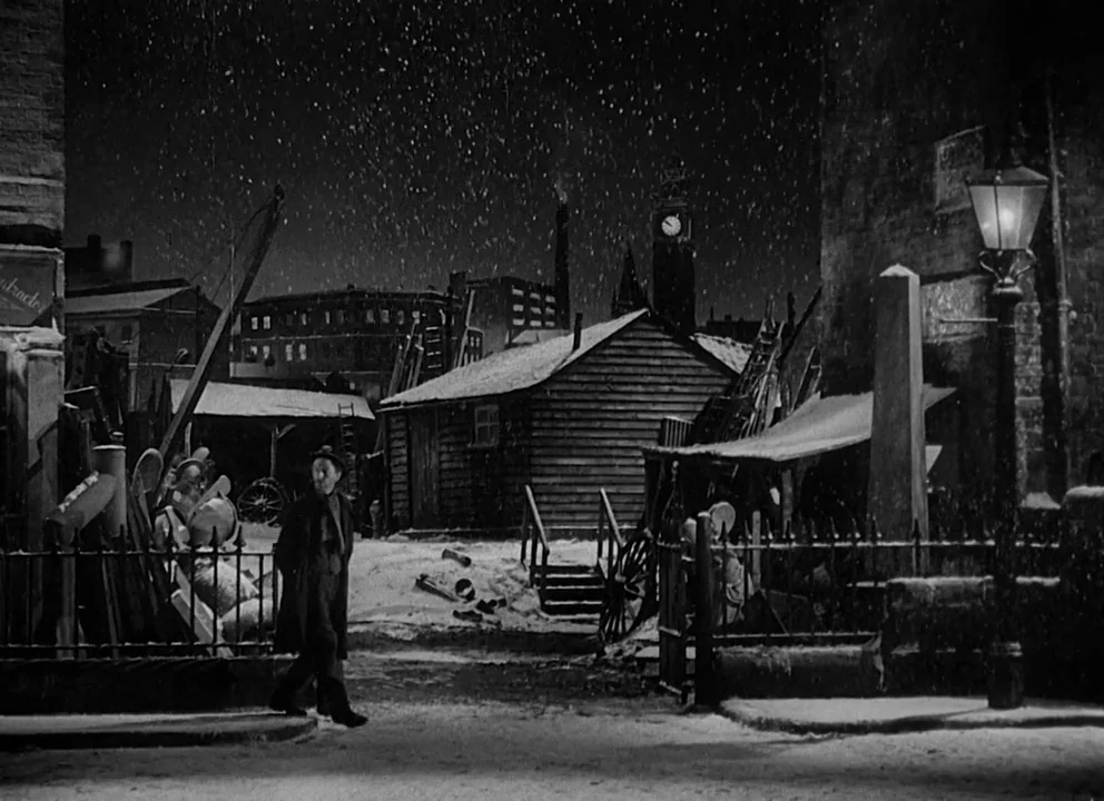 Odd Man Out (1947) movie still: wide — A cold, snow-covered industrial courtyard features a wooden shack and an old clock tower. A man in a dark…