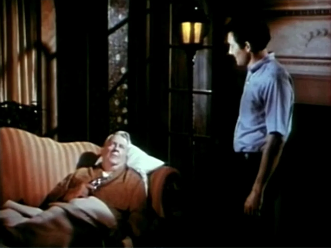 In the Year 2889 (1969) movie still: medium — A dark living room contains a striped upholstered sofa and a floor lamp. An older man resting on the…