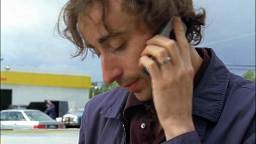 Old Joy (2006) movie still: closeup — A man stands outside a gas station on a gray, overcast day. The subject holds a cell phone to his ear with his…