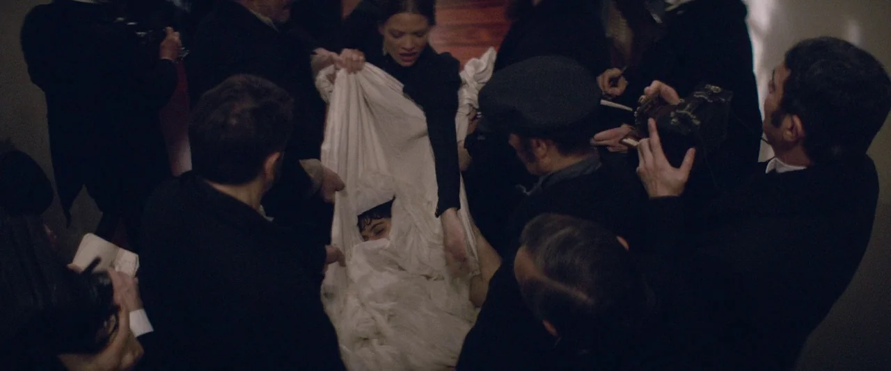 The Dancer movie still: medium — A dimly lit indoor space crowded with men in dark suits. A woman stands in the center wrapping a person in a white…
