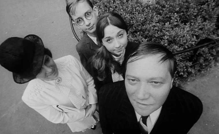 The Cremator (1969) movie still: medium — A group of four people stands outdoors near a dense evergreen hedge. They are arranged in a cluster and…
