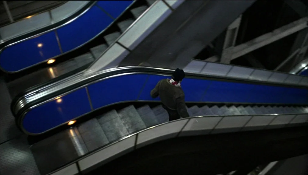 The American Friend (1977) movie still: wide — A dark subterranean transit area with intersecting metal escalators featuring bright blue side…
