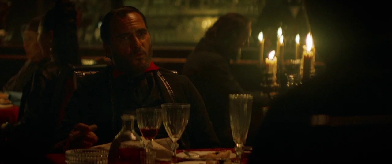 The Sisters Brothers (2018) movie still: medium — A dimly lit room filled with shadows and the warm flicker of candles on tables. A man sits at a…