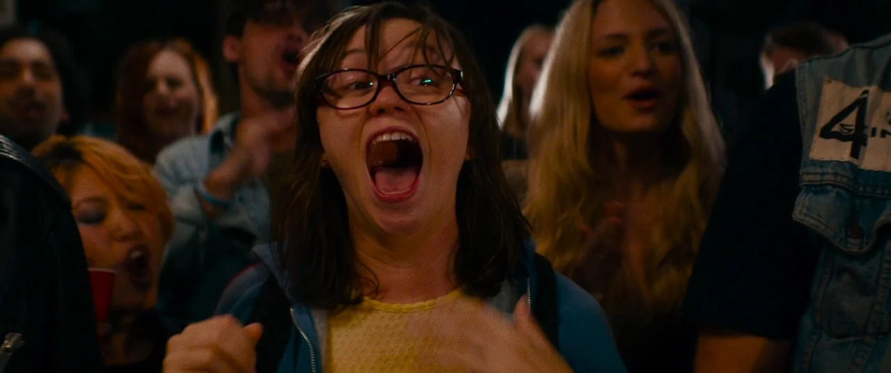 Dinner in America (2020) movie still: medium — A group of young adults are gathered in a dark, crowded indoor concert space. A woman with glasses in…
