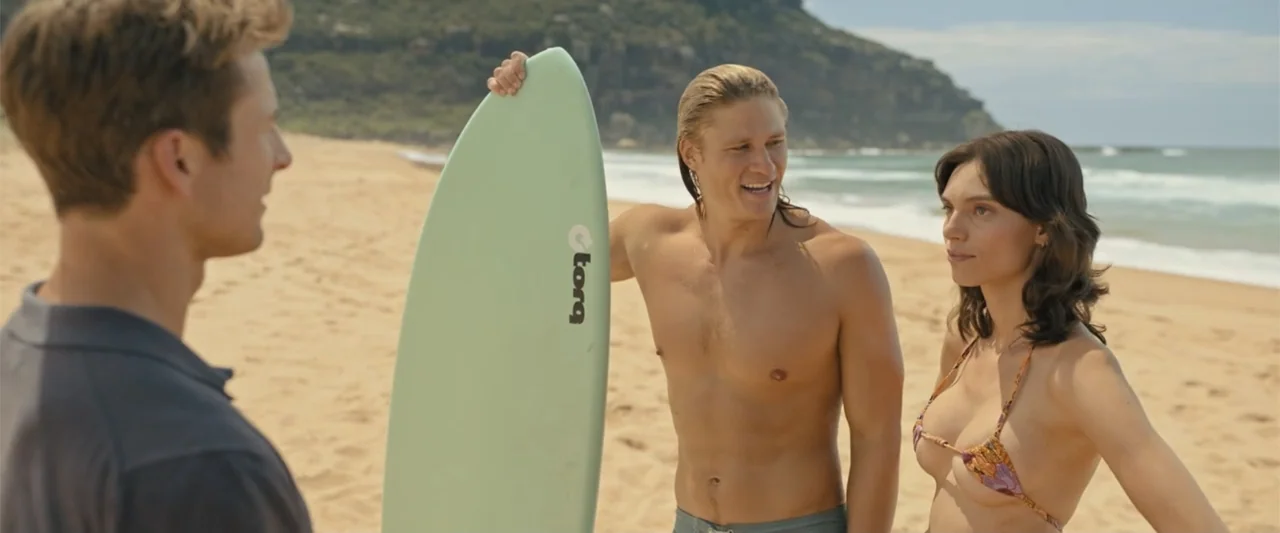 Anyone But You (2023) movie still: medium — A sunny sand beach with a cliff line in the background near the ocean. Three young adults stand together…