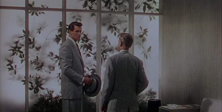 Magnificent Obsession (1954) movie still: medium — A modern room features a large window wall partially obscured by leafy outdoor plants. Two men in…