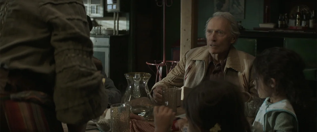 Cry Macho (2021) movie still: medium — A wooden dining area with green walls and a cluttered table containing a pitcher. An elderly man with grey…
