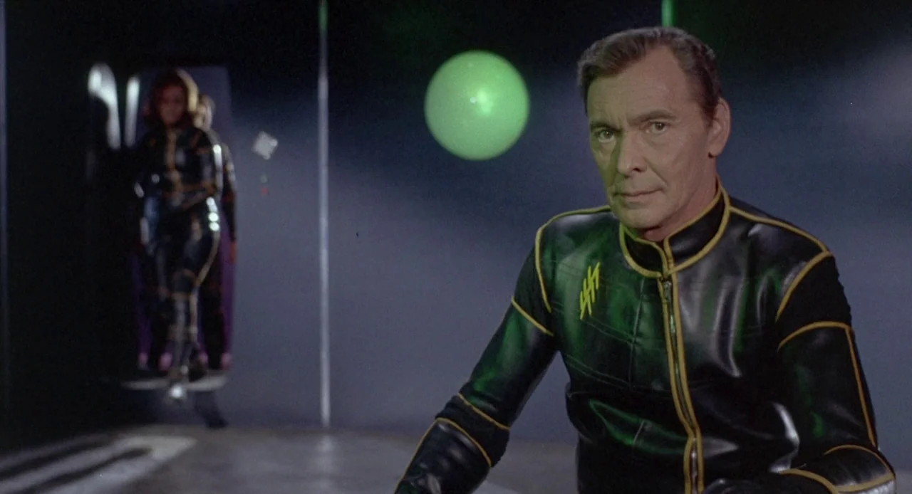 Planet of the Vampires (1965) movie still: medium — A dark, spartan interior with a prominent glowing green sphere floating in midair. A man in a…