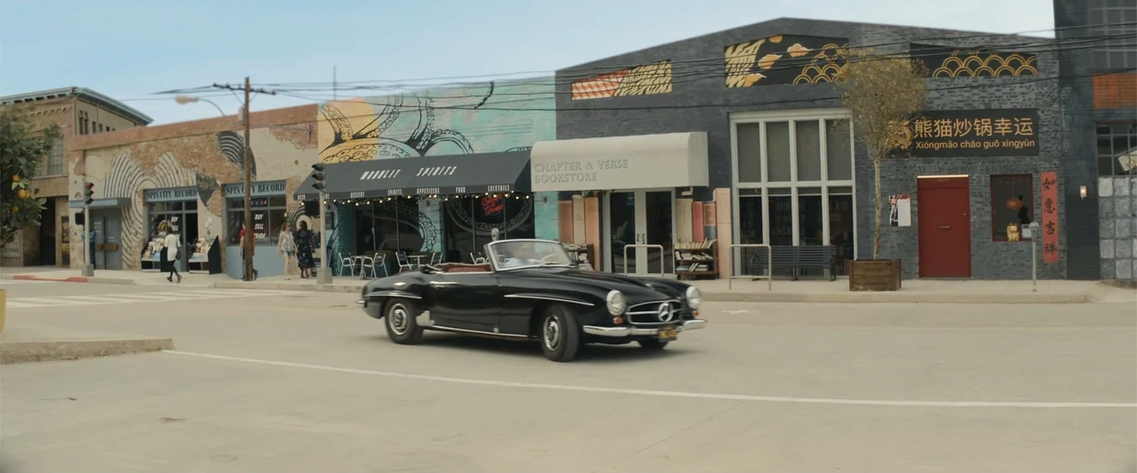 Maintenance Required (2025) movie still: wide — A suburban street lined with colorful storefronts features a mural and brick facades. A black…