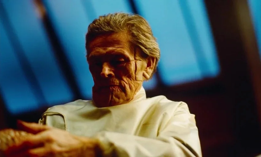 Poor Things (2023) movie still: closeup — A hospital room interior with large windows framing a blue background. An elderly man in a white medical…