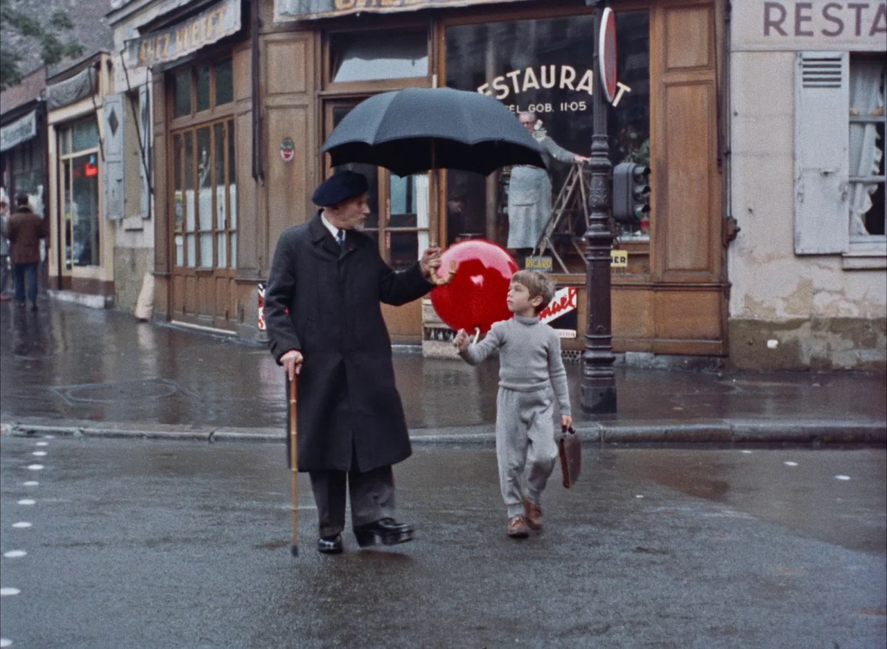 The Red Balloon (1956) movie still: fullbody — An urban intersection in Paris during a rainy day with storefronts in the background. An elderly man…