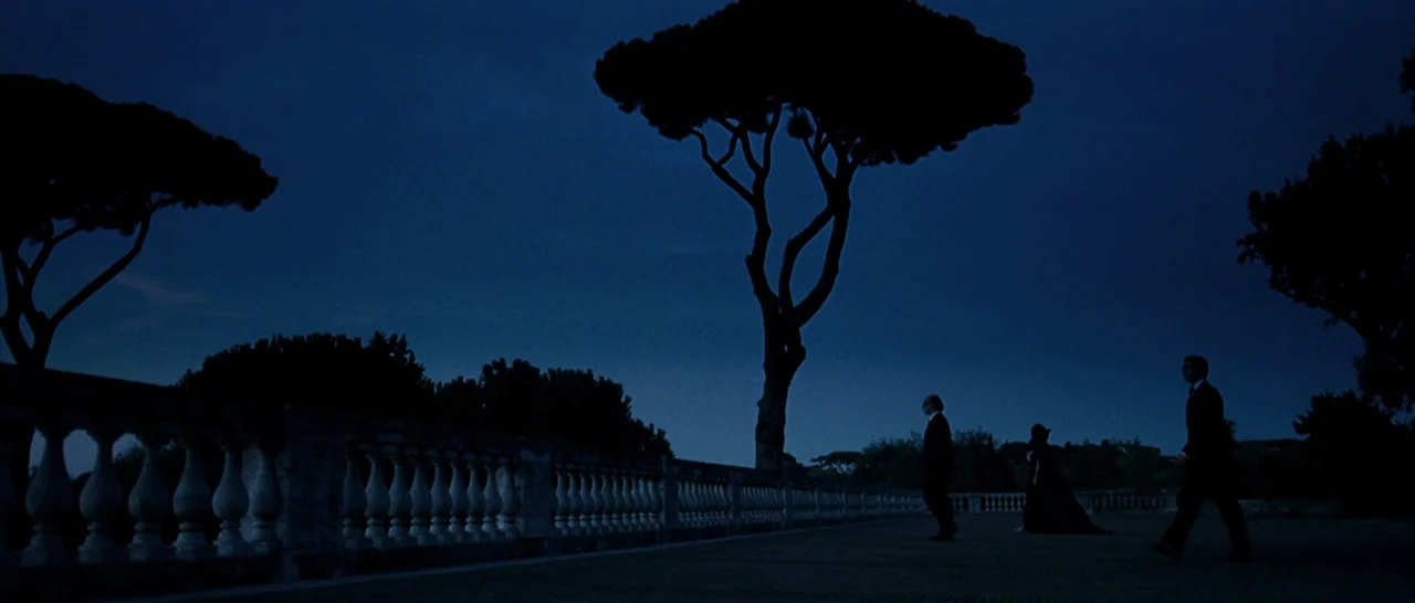 The Great Beauty (2013) movie still: wide — A stone terrace with a classical balustrade sits beneath large, dark umbrella pine trees. Three figures…