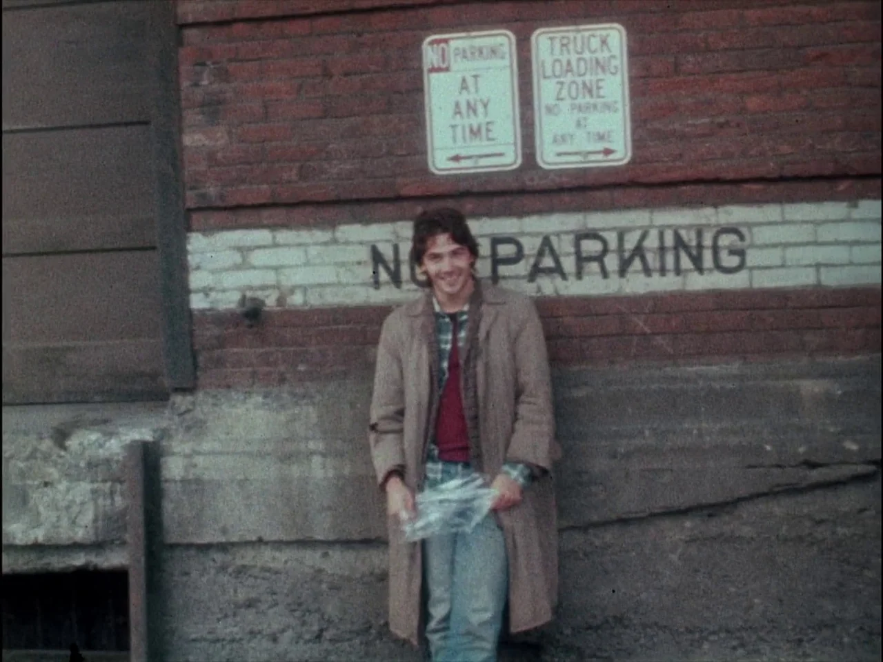 Mala Noche (1986) movie still: fullbody — A man stands in front of a red brick wall featuring large no-parking signage. He holds a clear plastic bag…