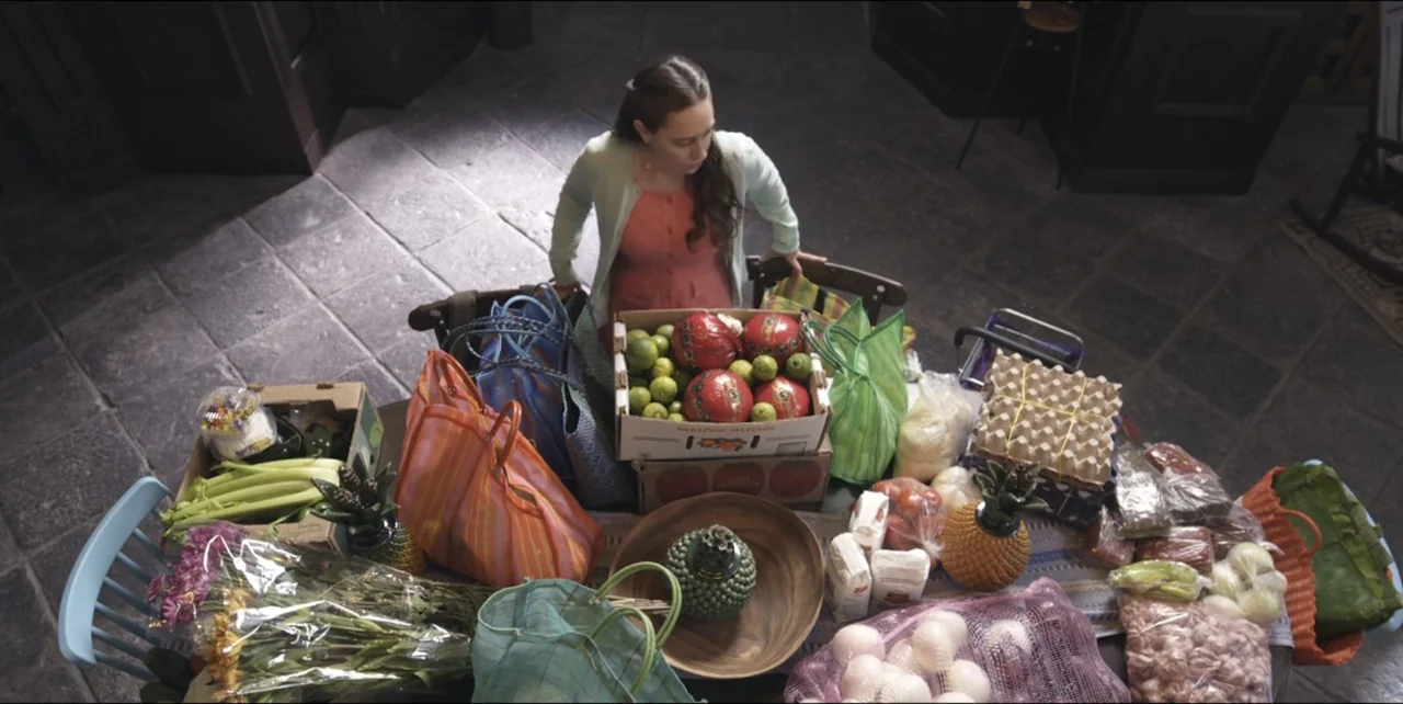El sabor de la Navidad (2023) movie still: medium — A stone-floored kitchen features a large wooden table covered in diverse groceries and bags. A…