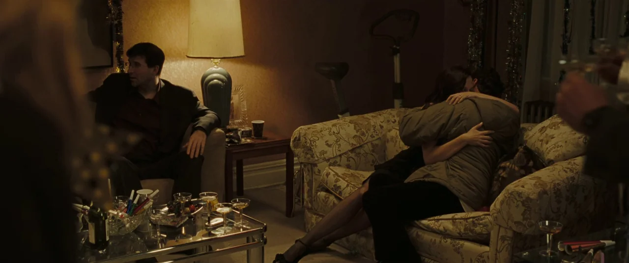Two Lovers (2008) movie still: medium — A dimly lit residential space contains a patterned fabric couch and glass-topped coffee table. Two people…