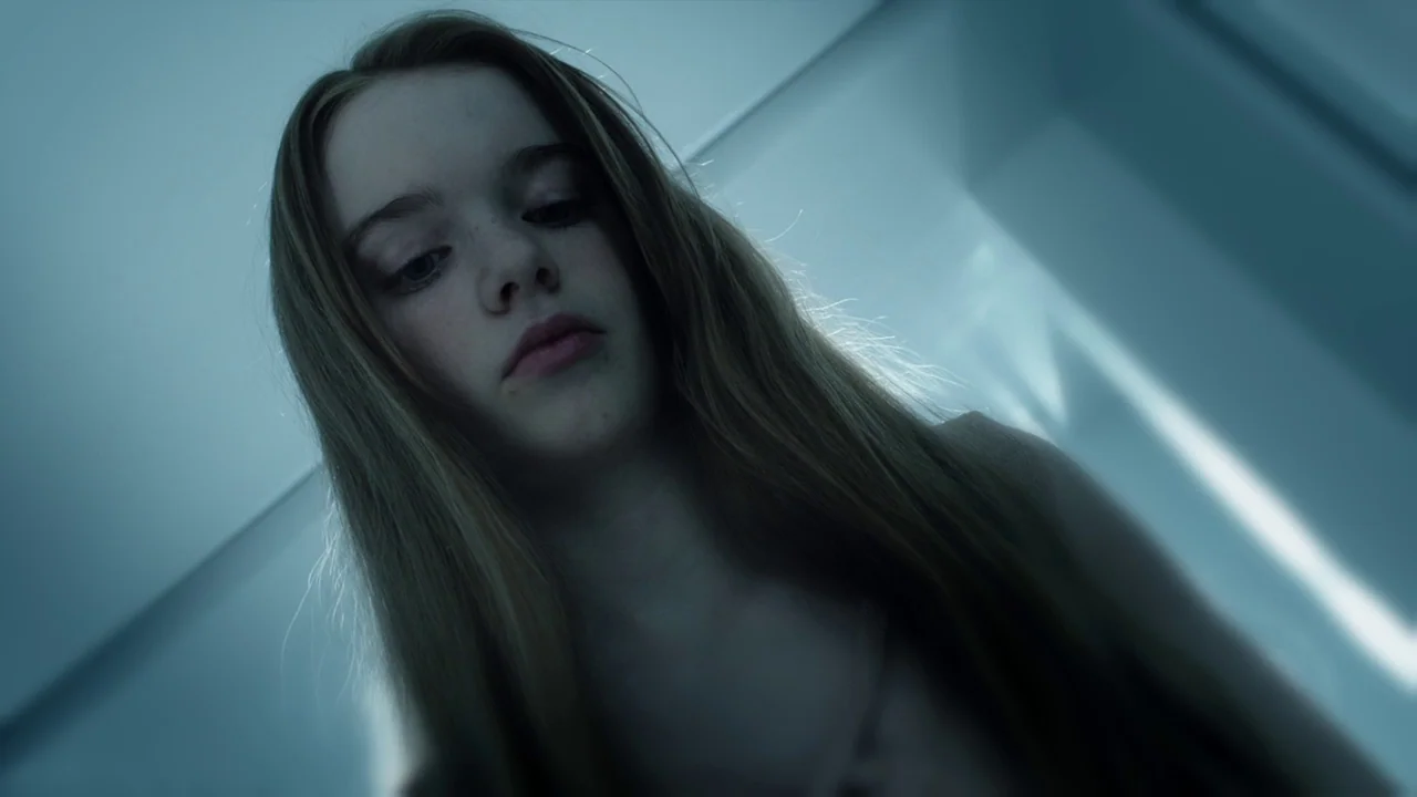 Cult of Chucky (2017) movie still: closeup — A teenager stands in a light-filled stairwell with white walls and sharp angles. She looks directly…