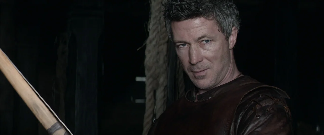 King Arthur: Legend of the Sword (2017) movie still: closeup — A dimly lit space with thick rope hanging in the dark background. A man wears leather…