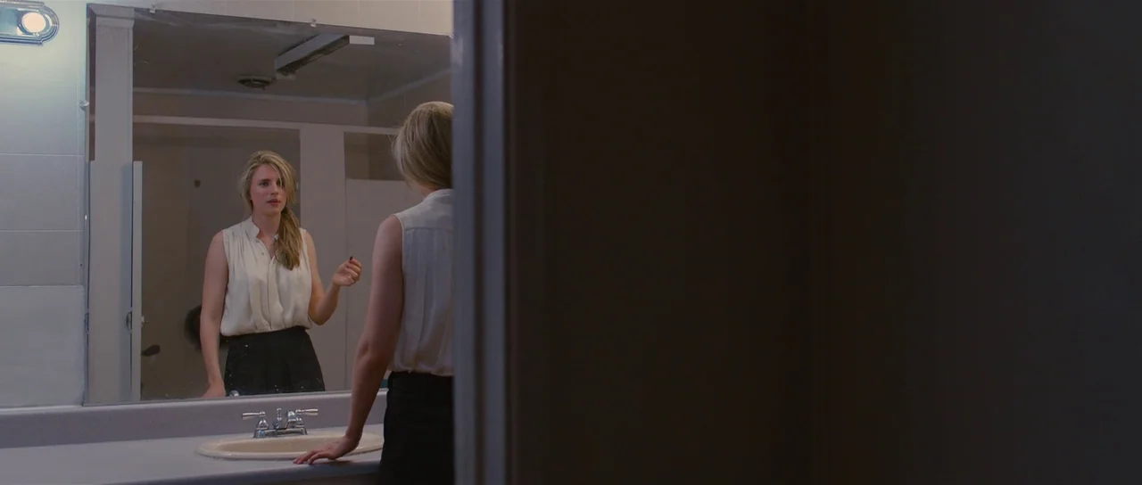 The East (2013) movie still: medium — A public restroom with white tiled walls and a large vanity mirror. The subject stands before the mirror while…