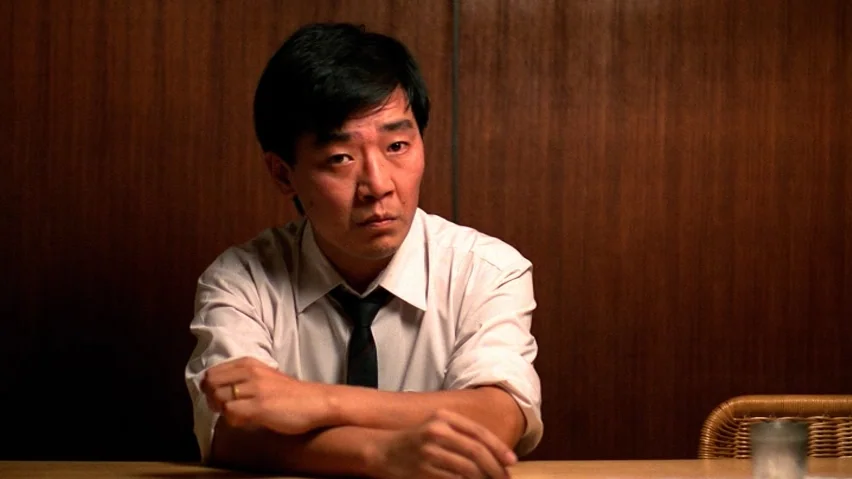 The Terrorizers (1986) movie still: medium — A man sits at a table in front of a dark wooden paneled wall. He leans forward with his arms crossed on…