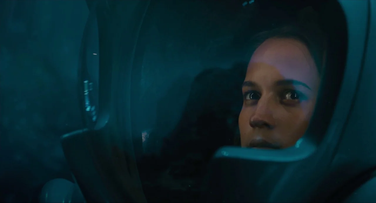 Submergence movie still: closeup — A close view through the glass faceplate of a diving helmet. The woman stares forward into the dark blue…
