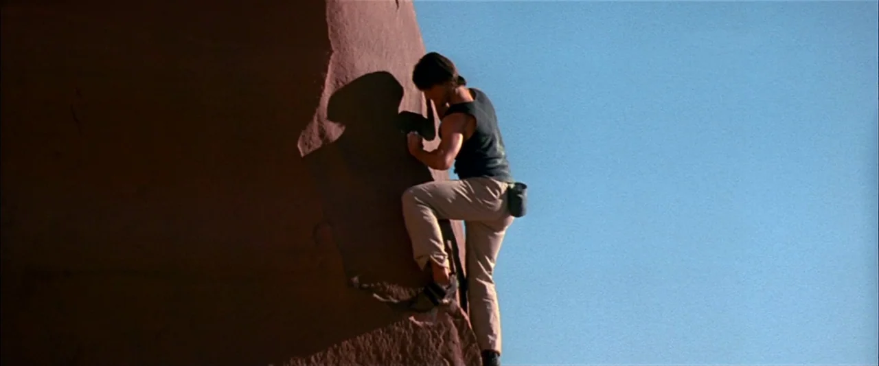 Mission: Impossible II (2000) movie still: fullbody — A climber scales the edge of a massive, sunlit sandstone rock formation. A clear bright blue…