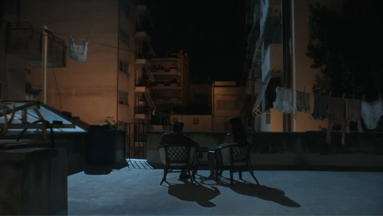 Hot Milk (2025) movie still: wide — A concrete rooftop terrace is surrounded by multi-story residential apartment blocks at night. A man and a woman…
