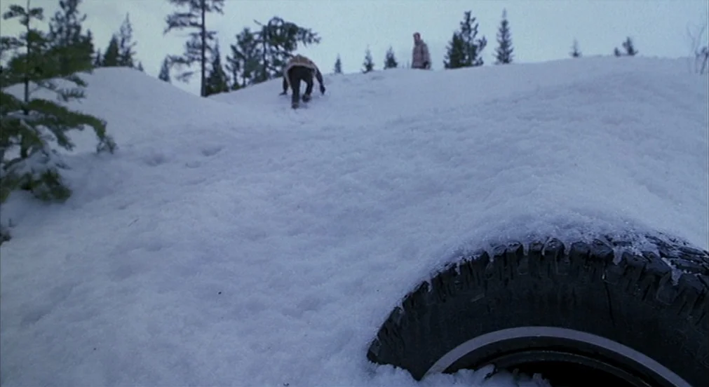 Misery (1990) movie still: wide — A snow-covered incline with a single vehicle tire protruding from the foreground drift. Two men navigate the…