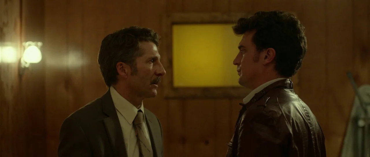 Faults (2014) movie still: medium — Two men in a wood-paneled room face one another in a medium profile shot. The older man in a suit looks at the…