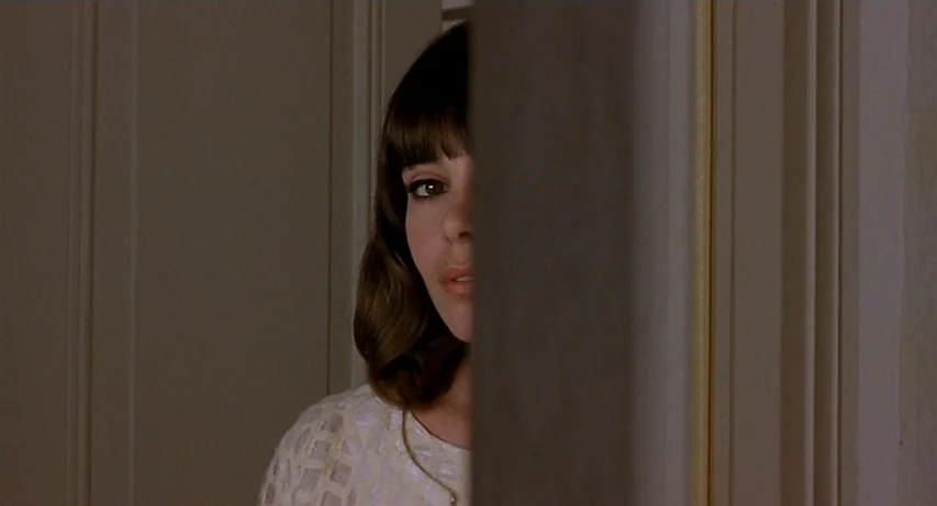 CQ (2001) movie still: closeup — A woman looks through a narrow opening between a wall and a door frame. The surrounding environment consists of…