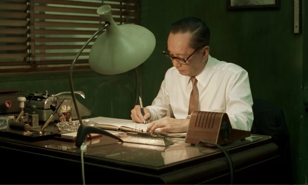 In the Mood for Love (2000) movie still: medium — A dimly lit office features an older man sitting at a dark wooden desk. He uses a pen to write in…