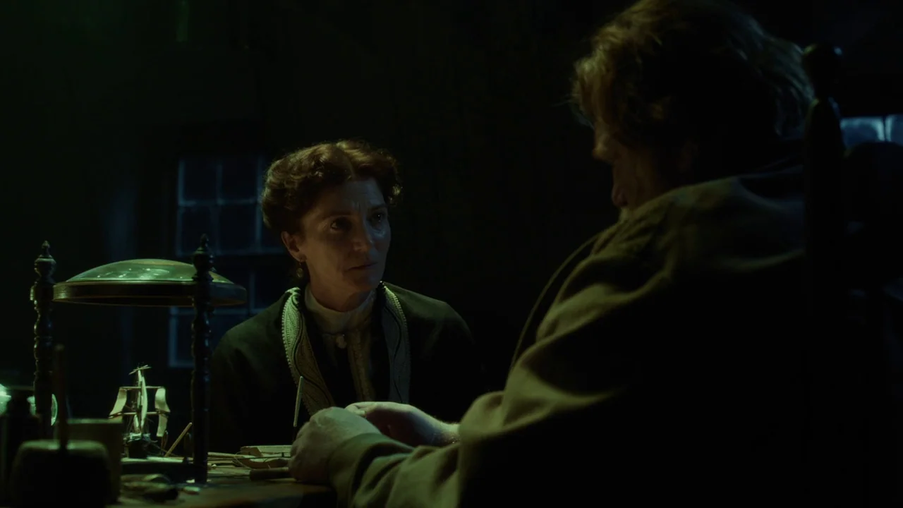 In The Heart of the Sea (2014) movie still: medium — A dimly lit room contains a cluttered wooden desk with an antique model ship. A woman watches a…