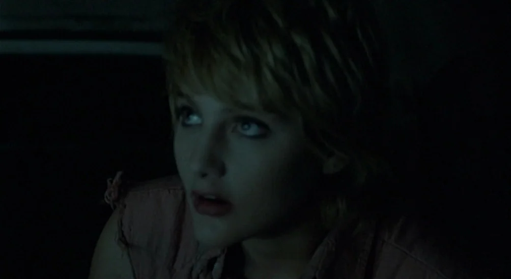 Near Dark (1987) movie still: closeup — A dark, dimly lit interior space with shadows obscuring the surroundings. A young woman with short blonde…