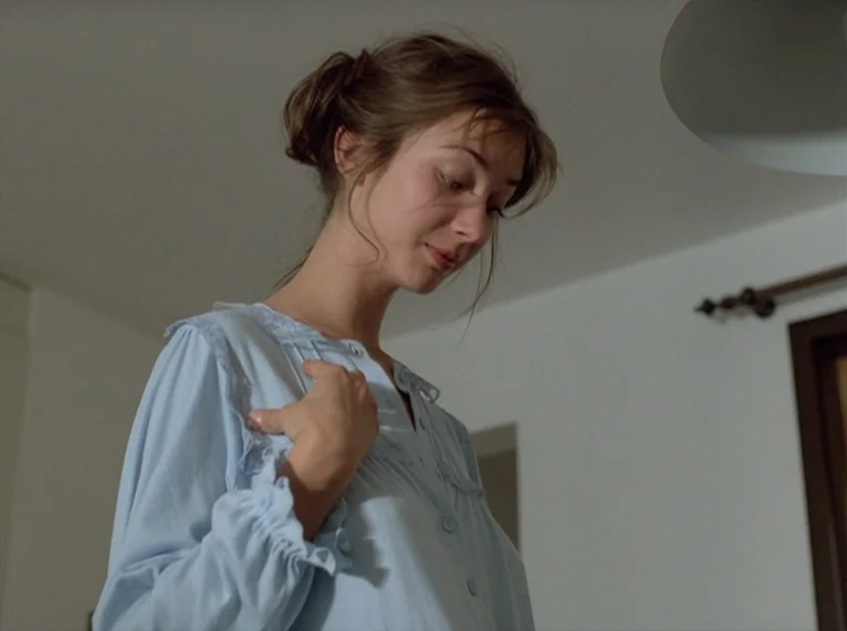 Hail Mary (1985) movie still: medium — A woman stands in a bright room with plain white walls. She gazes downward while wearing a blue nightgown…