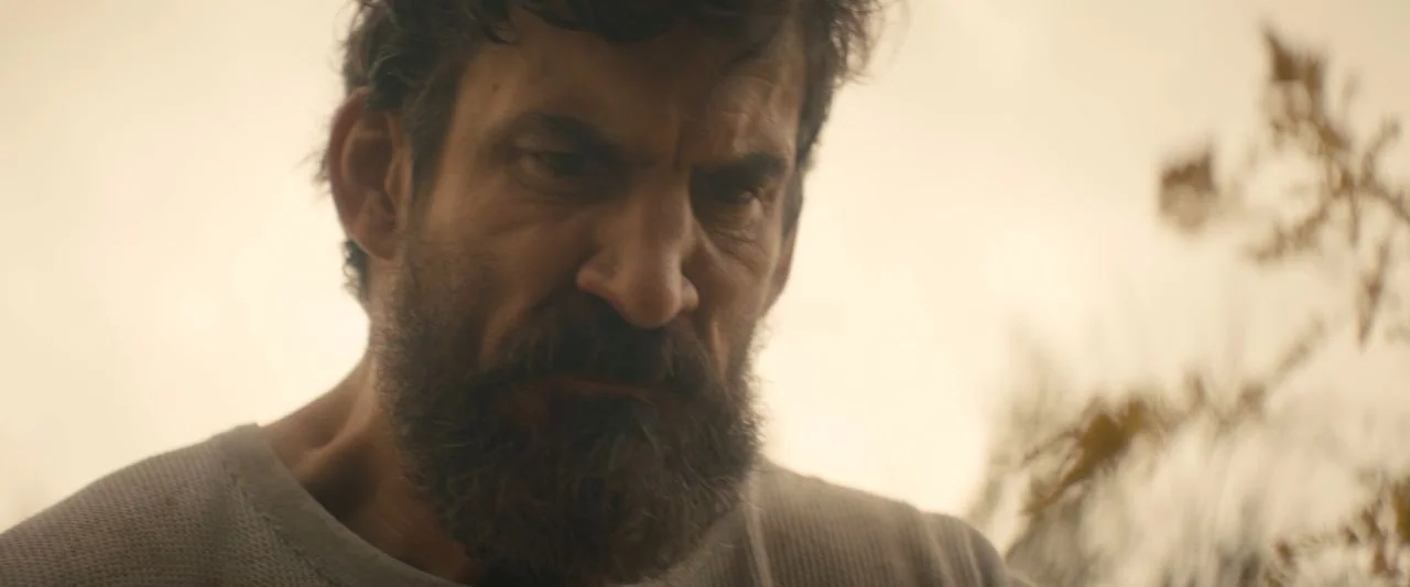 Becky (2020) movie still: closeup — A man stands outdoors in a field with dry vegetation visible to the side. The man looks down with his brow…